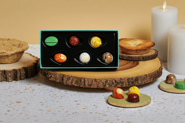 Box of assorted artisan chocolates displayed on a wooden serving board with candles in the background.