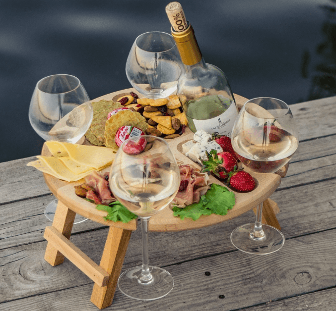 Outdoor wine and charcuterie setup with four wine glasses on a small wooden picnic table by the water.